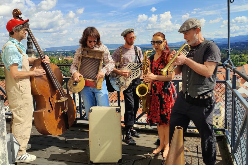 Musizierende der Band Los Boozan Dukes stehen mit ihren Instrumenten auf dem Außenbereich des Johannisturms und spielen Musik im Sonnenschein. Im Hintergrund die Ausläufer des Zittauer Gebirges.