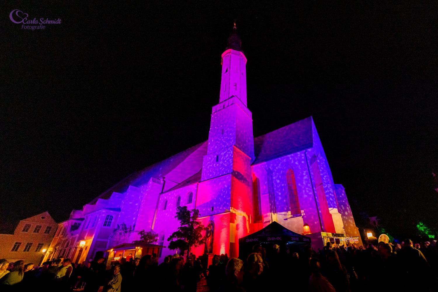 Foto klášterního kostela. Celá budova je až po vrchol věže osvícena různými červenými a fialovými odstíny. V popředí stojí početné množství lidí.