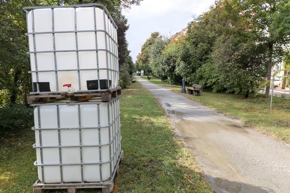 Zwei schwere Wasserbehälter auf Paletten stehen übereinander gestapelt am Rand eines Gehwegs in einer Parkanlage