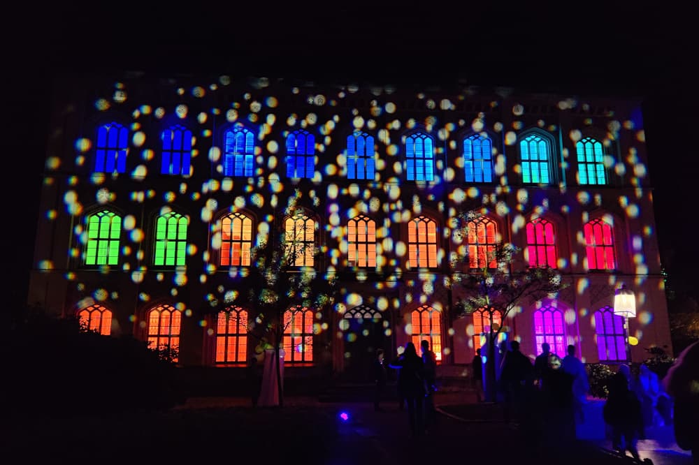 Foto der Baugewerkeschule bei Nacht. Das Gebäude ist im Inneren mit unterschiedlichen farbigen Lichtern beleuchtet und von Außen mit weißen Punkten angestrahlt.